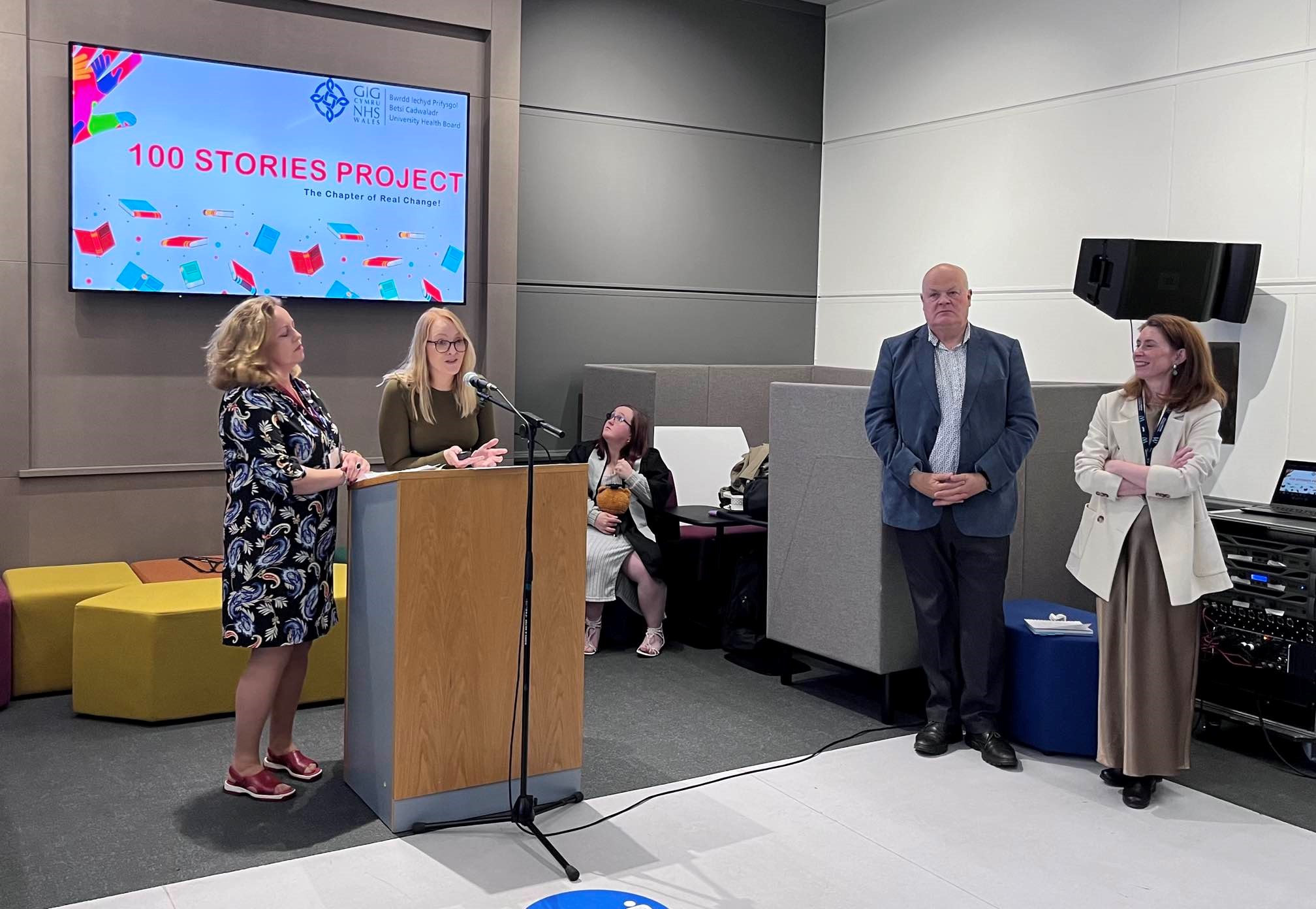 100 Stories Project Celebration Event. Keynote speakers standing at a lectern with people listening and smiling.