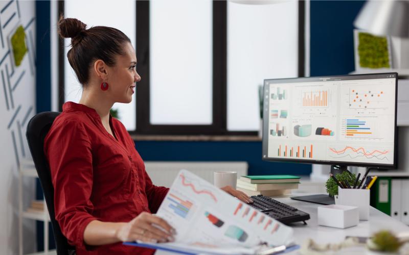 Woman sat at computer with papers including graphs, bar charts and tables