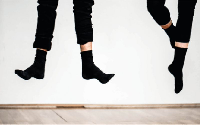 Photograph of two pairs of feet in black socks and black trousers, lifted from the ground and dancing