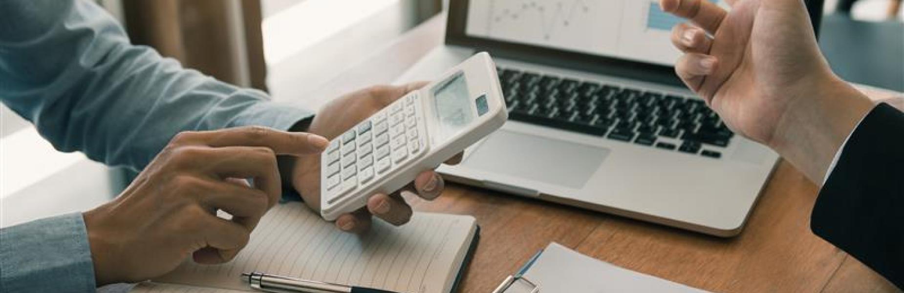 Two people discussing data at a table with a laptop, calculator, and documents