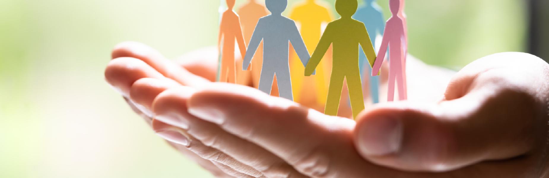 Close-up of two hands gently cupping a group of colorful paper cutouts shaped like human figures.