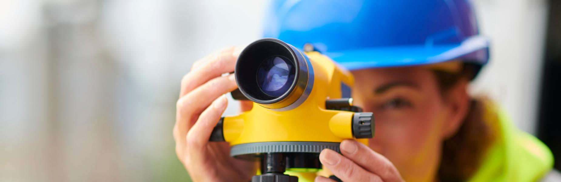 Mae gweithwr adeiladu mewn het galed las yn defnyddio offerynnau arolygu wedi'u mowntio ar drycedd mewn safle gwaith.