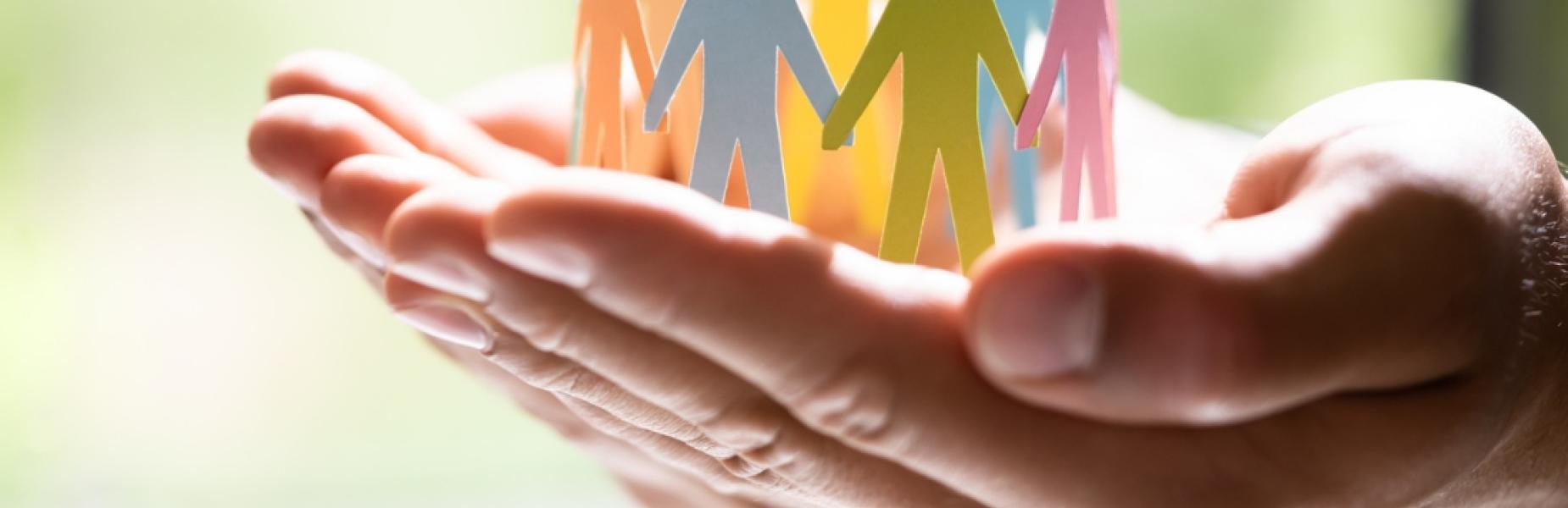 Grŵp o ffigurau papur lliwgar o bobl yn sefyll gyda’i gilydd mewn par o ddwylo ar agor, yn cynrychioli cymuned, cefnogaeth a chynhwysiant.