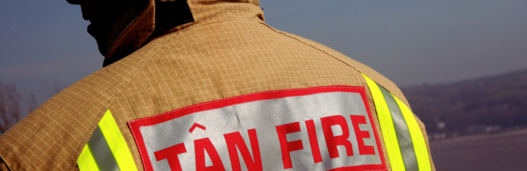 Firefighter wearing a tan protective jacket and white helmet, viewed from behind, with a reflective ‘TÂN FIRE’ patch and high-visibility yellow strips on the back, standing outdoors against a coastal landscape and clear sky.
