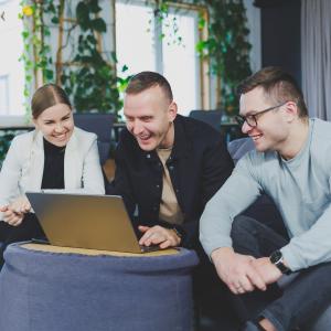 two men and a woman sat in a café on a sofa around a laptop smiling 